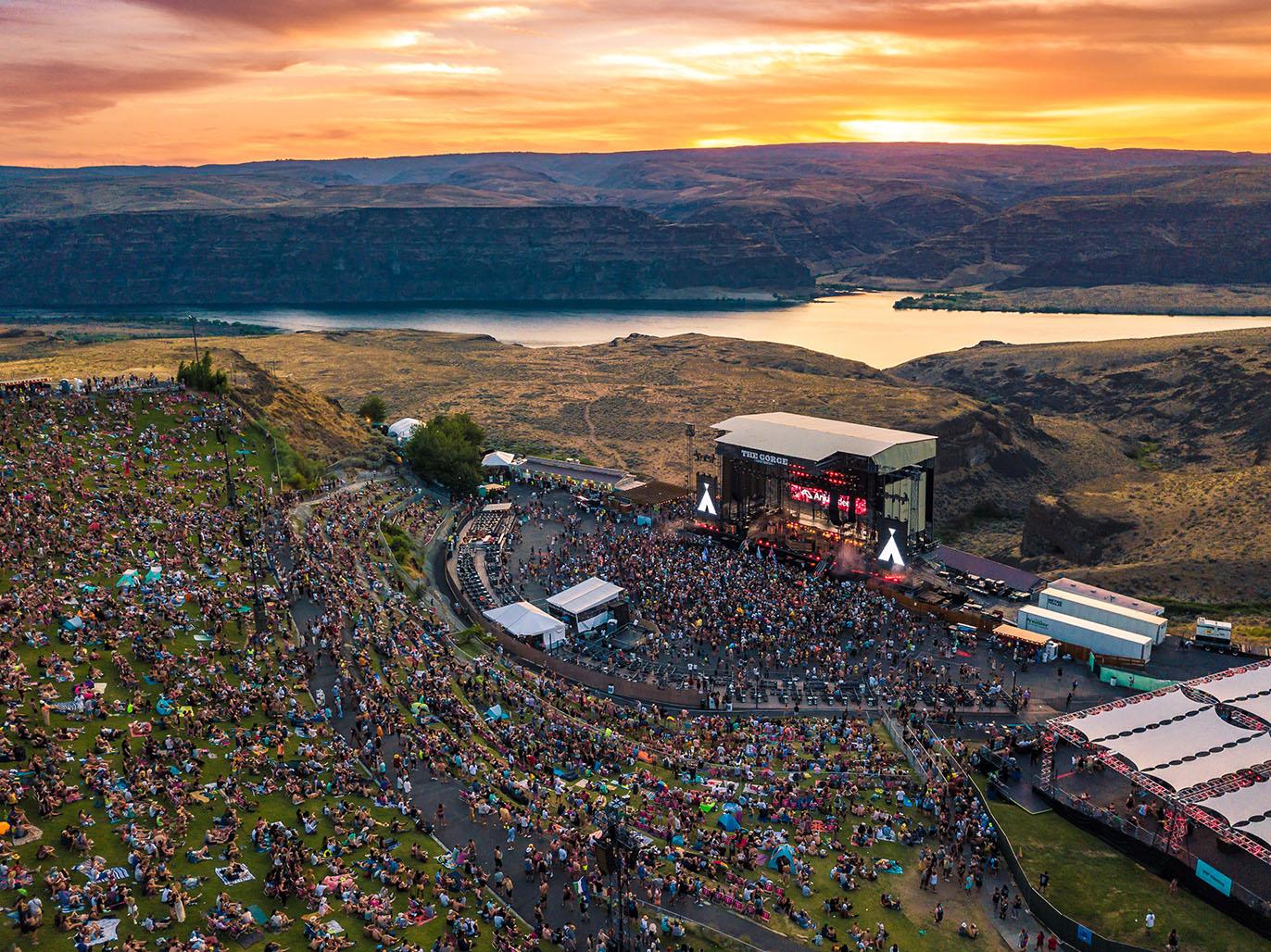 Festival-goers gathered at The Gorge Amphitheatre at dusk, with the stage lights beginning to glow against a stunning canyon backdrop and sunset reflection on the river.